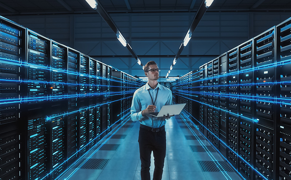 サーバーが並ぶ部屋でノートPCを持つ男性開発者。