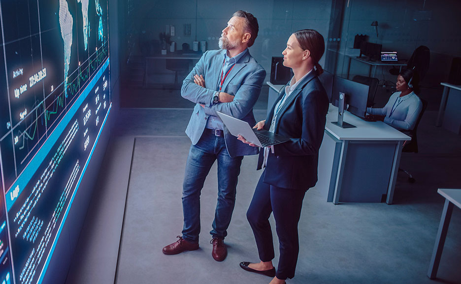 Two business colleagues in control room visualizing data in a big screen