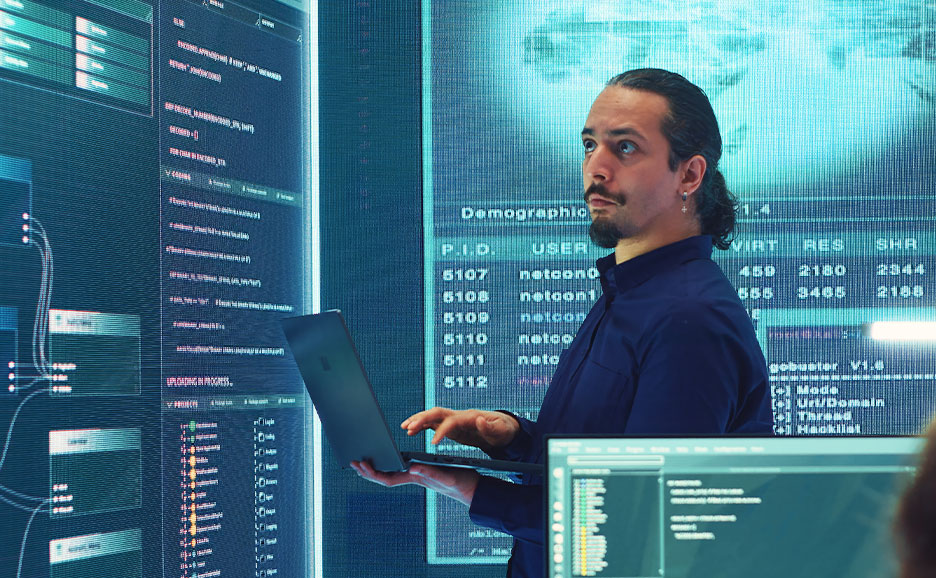 IT professional working on a laptop in front of large screens displaying integration software dashboards, code, and user data for managing complex workflows