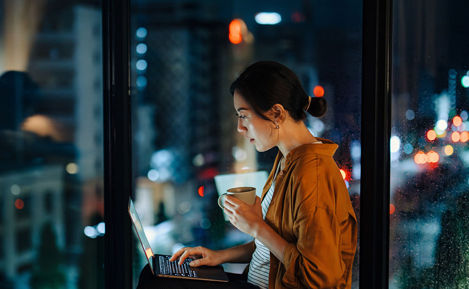 A woman using her laptop sitting by a window with a city lit up at night in the background