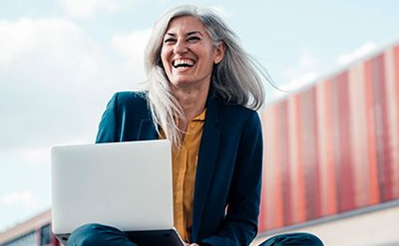A woman seated on the ground, working intently on her laptop, with a natural backdrop of grass and trees.