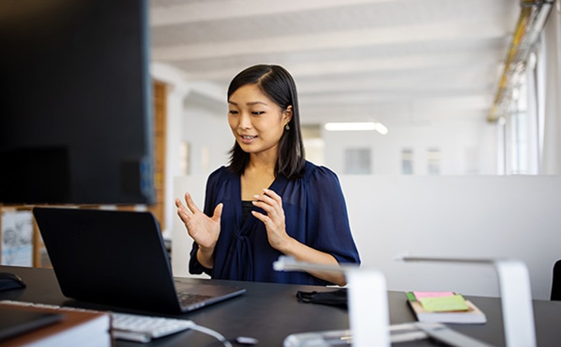 A woman sits at a desk, focused on her laptop, with a notepad and pen beside her.