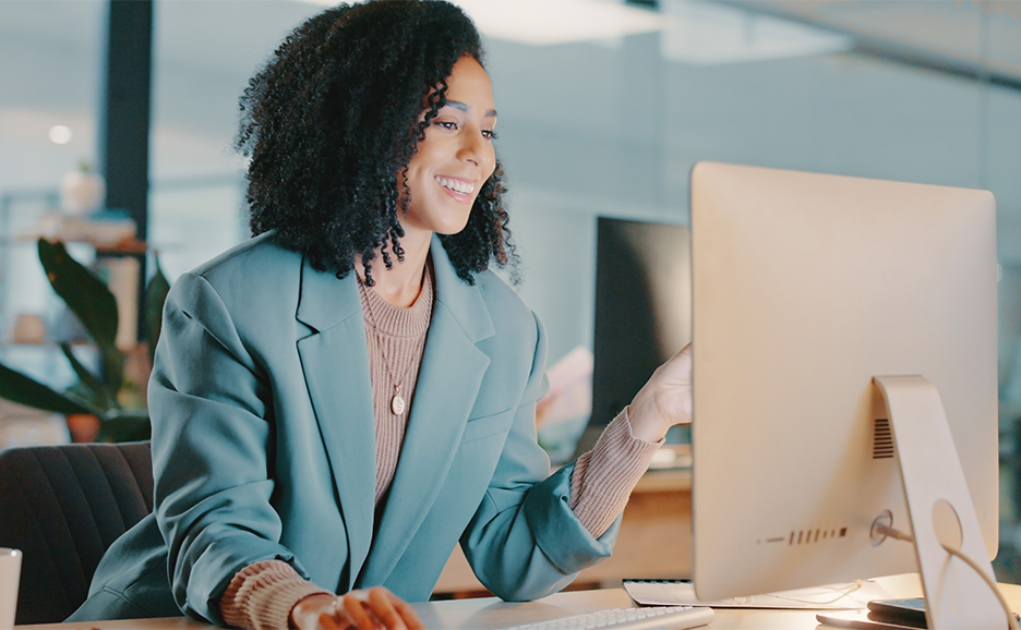 A woman is sitting at a desk, smiling while looking at a computer screen.