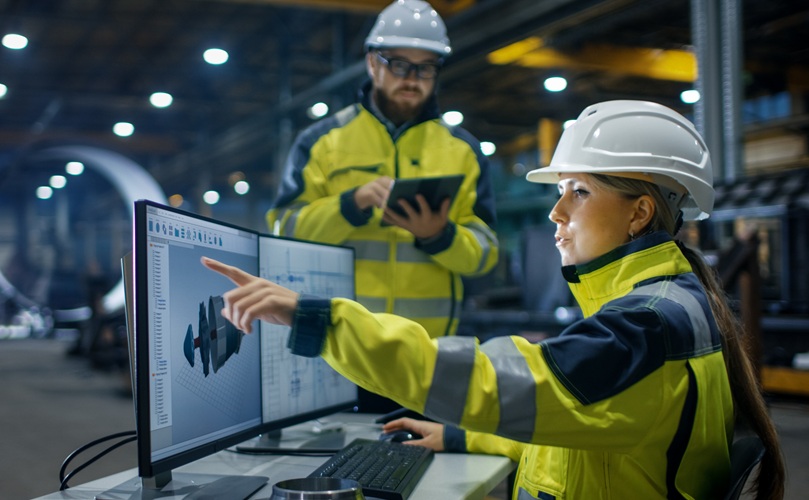 A female engineer in high-vis gear points at a computer screen displaying technical designs, while a male colleague with a tablet stands behind her.