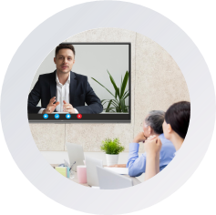 icon A diverse group of people seated in a conference room, engaged in discussion with a video screen displaying information.