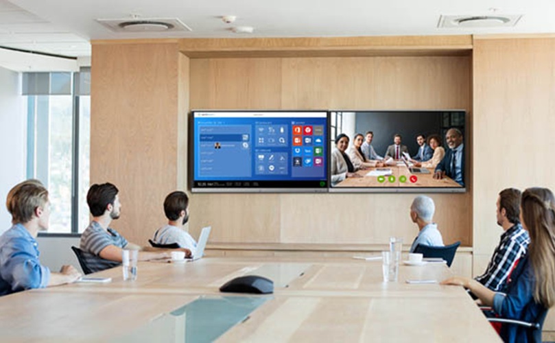 A group of professionals seated around a conference table, with two large screens displaying information in the background.