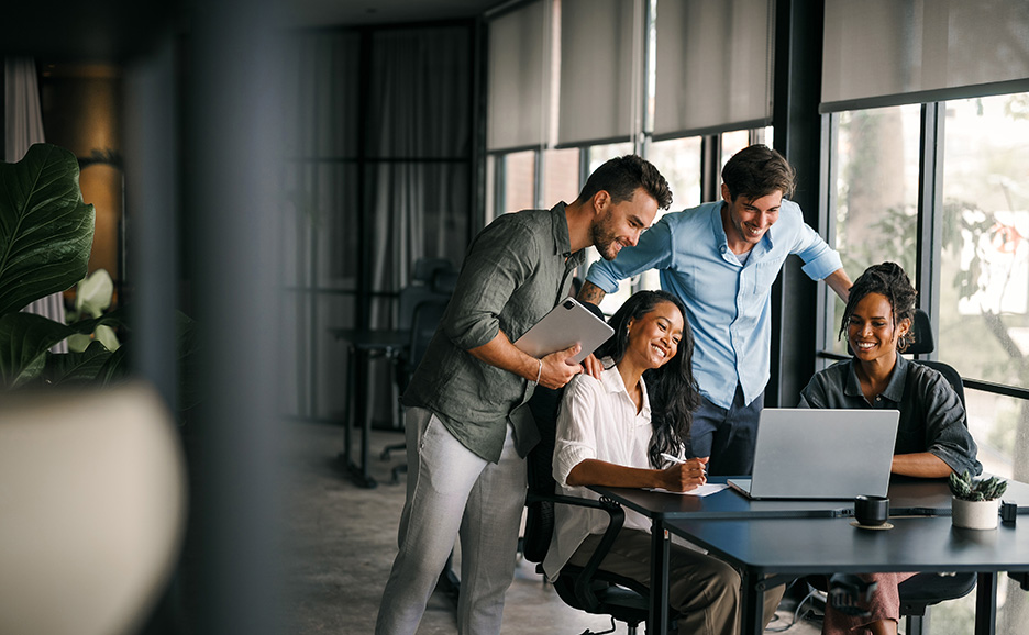 オフィスで、2人の男性が座っている女性たちとラップトップ画面を見て笑っています。