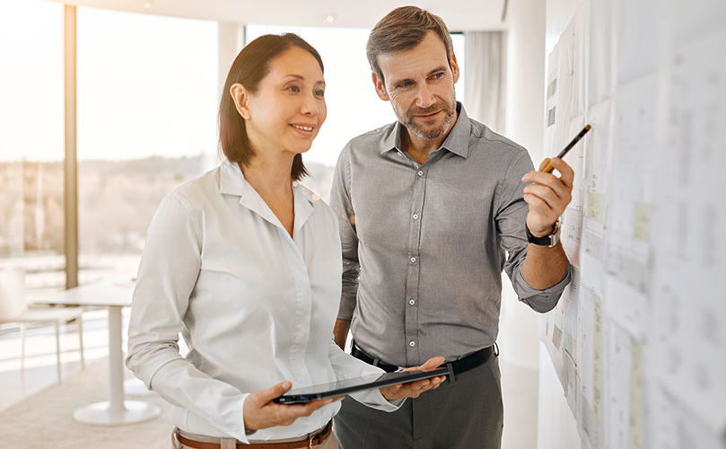A man and woman are positioned in front of a whiteboard, likely discussing or explaining information displayed.