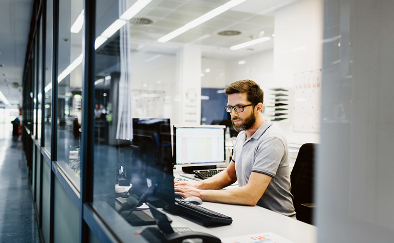 A man in glasses is diligently working on a computer, looking at the screen and typing with intent.