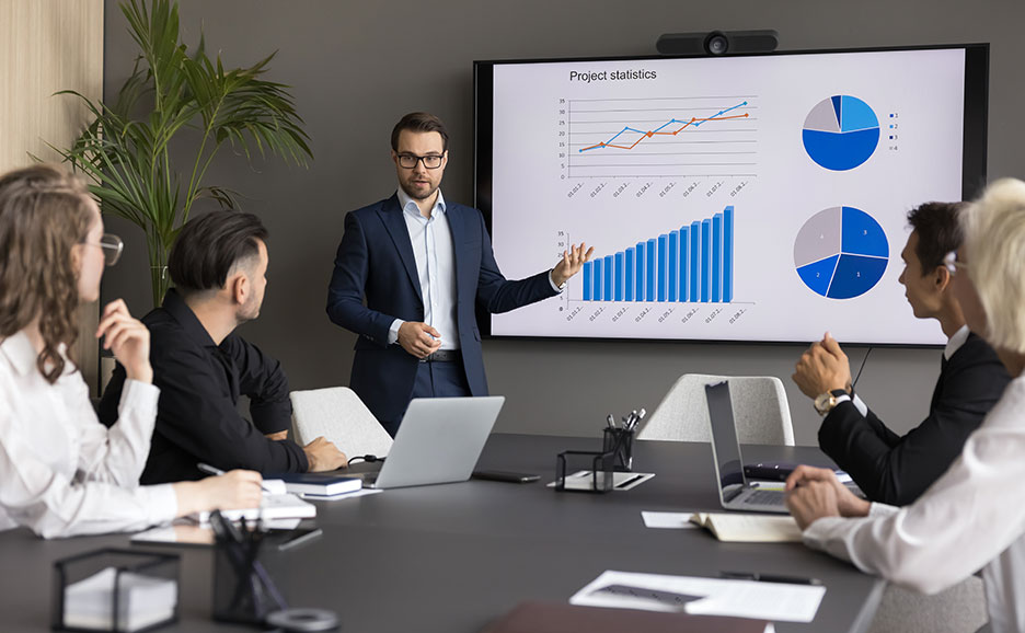 大型モニタに投影したグラフを説明している男性と聞いている4名の男女