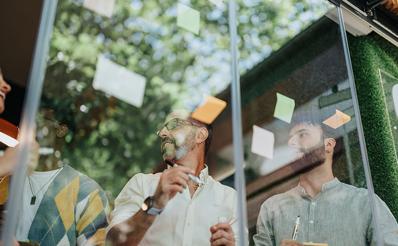 3人の男性がガラスのホワイトボードに貼られた付箋を見ながら話し合っている。