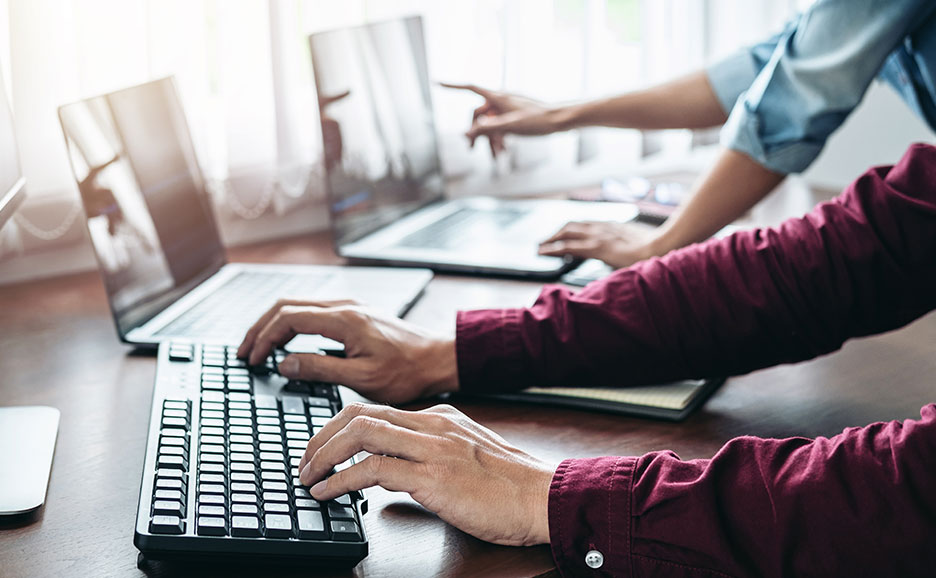Two pairs of hands working on PCs: one typing, one pointing.