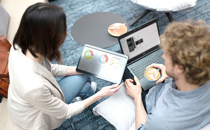 Two people sitting with laptops and coffee, one showing charts on a tablet