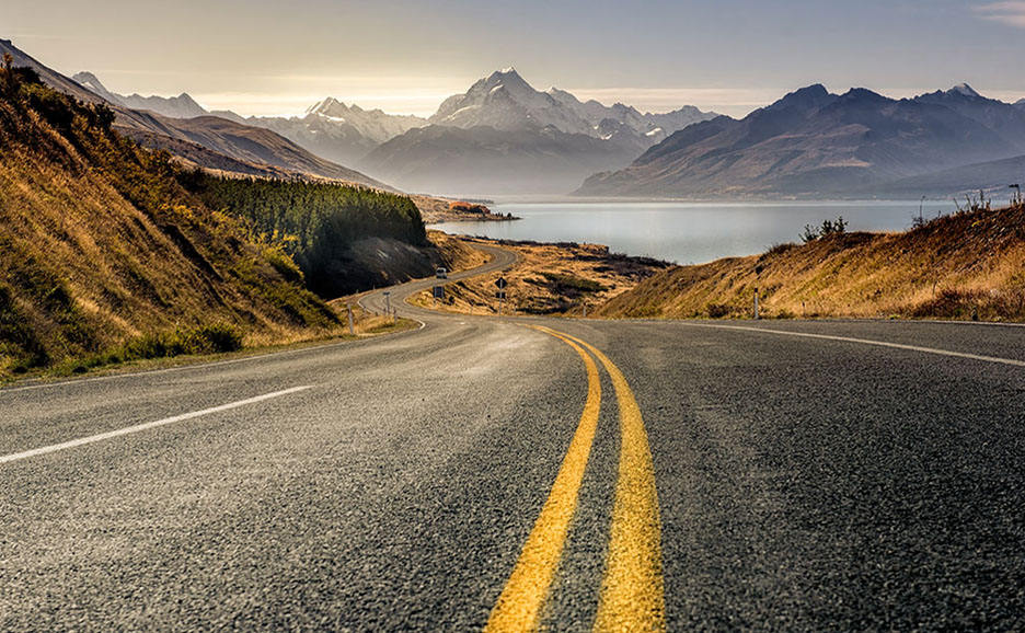 A road curving along a lakeside with snow-capped mountains in the background