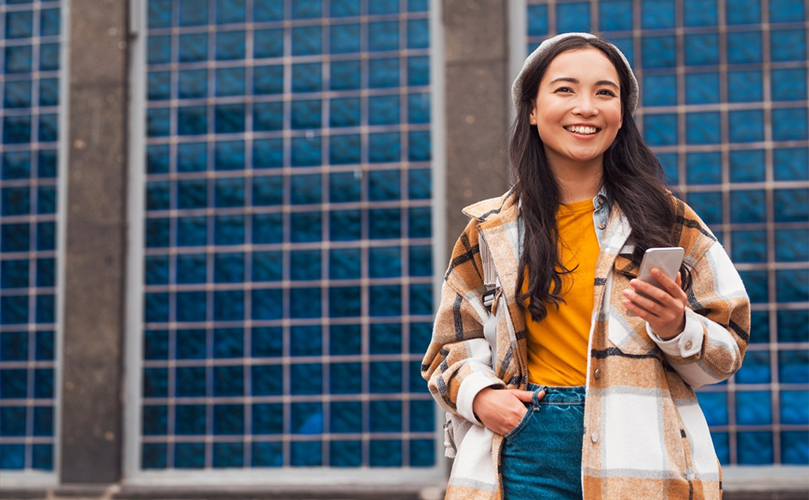 A woman wearing a plaid coat and jeans is holding her phone