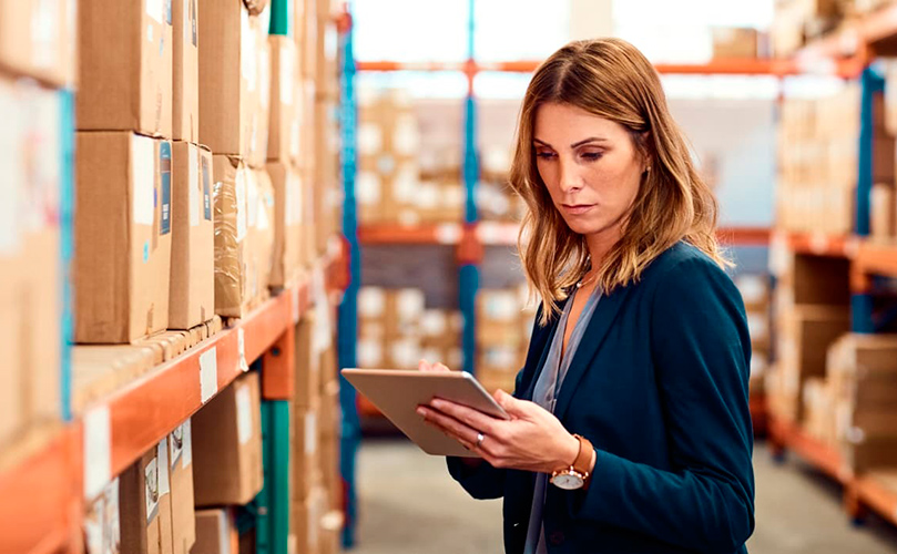 Photograph of a woman using a tablet to check inventory in a warehouse aisle filled with stacked cardboard boxes on metal shelves.