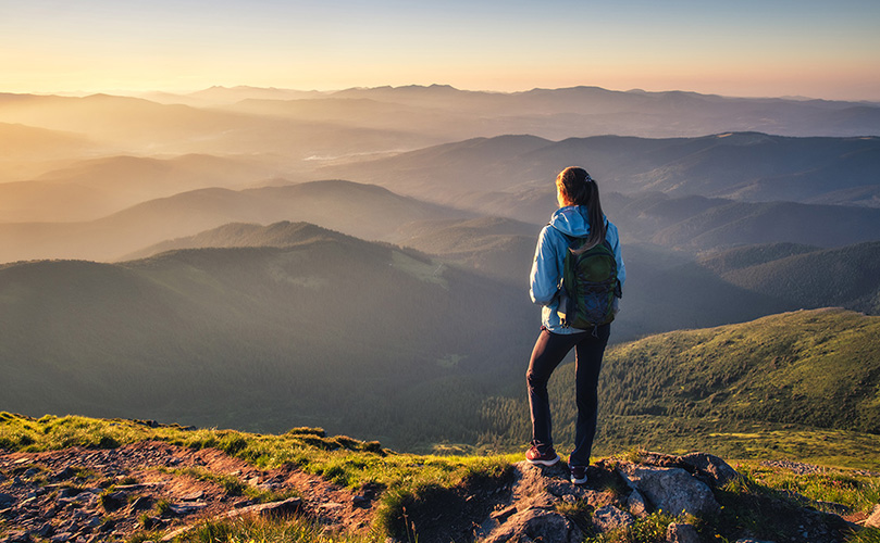 Eine Frau steht auf einer Felsformation und betrachtet die Berge bei Sonnenaufgang
