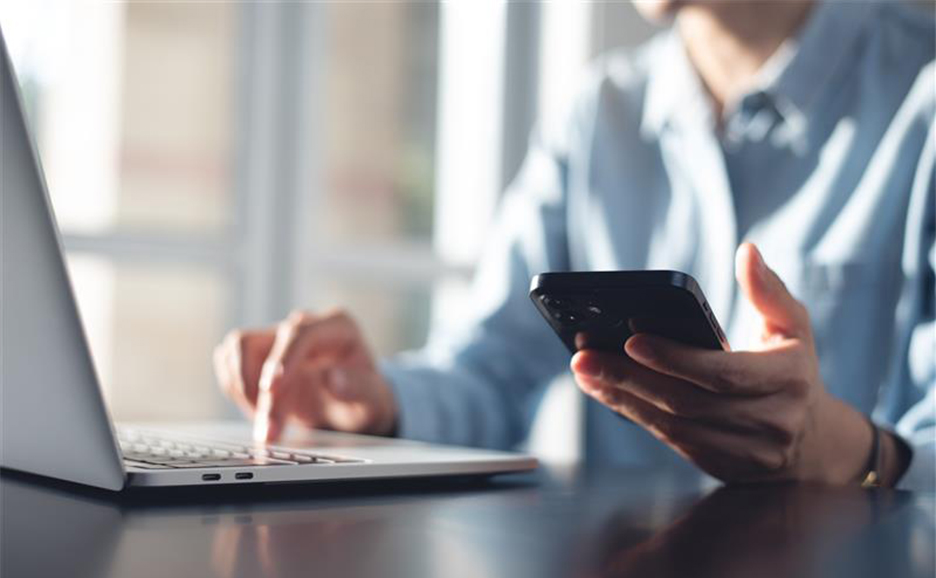 ノートPCを操作しながらスマートフォンを持つ人の手元のクローズアップ。