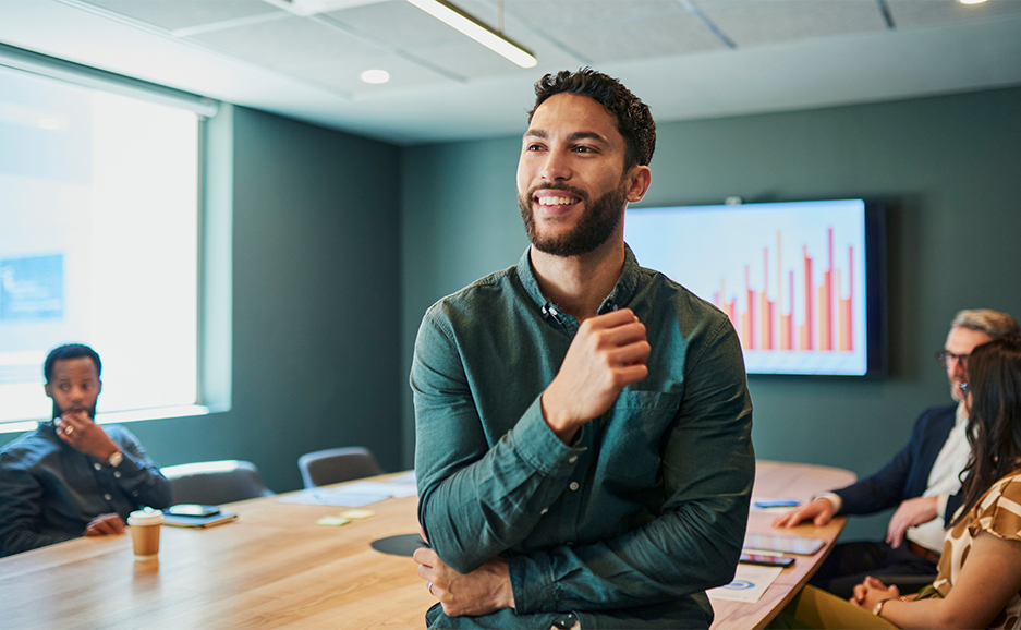 会議室で笑顔で前方を見るビジネスパーソンと、背景に会議の参加者とモニターにグラフが映る様子。