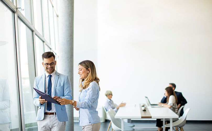 笑顔の男女が書類を見ている。奥では会議が行われている。
