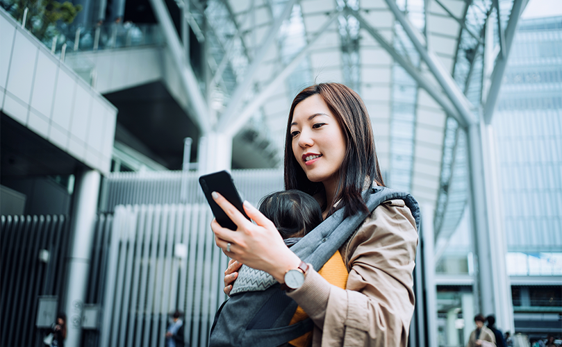 母親が赤ん坊を抱えながら、モダンな建物の外でスマートフォンを使っています。