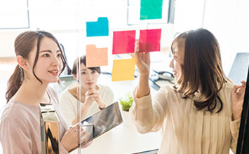 会議室で、女性たちがガラスのボードに付箋を貼る様子。