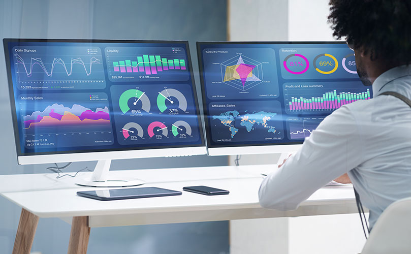 男性が机に座り、2つのモニターに数種類のチャートやグラフのデータダッシュボードが表示されている