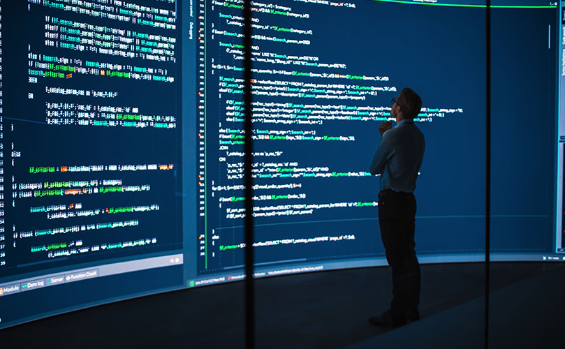 コードが映る巨大スクリーンを見つめ熟考する技術者