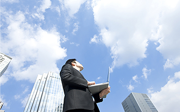 ビル街,ノートPCを手に空を見上げる男性