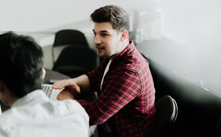 職場で一人がノートパソコンを操作し、もう一人が話している姿。