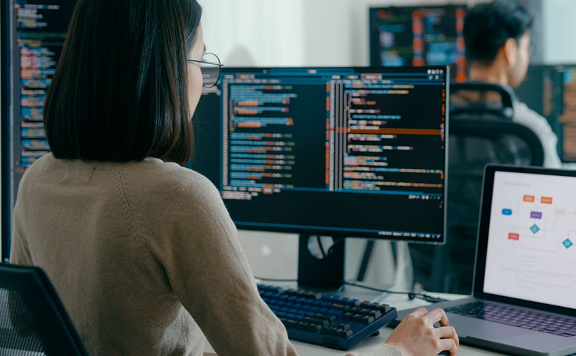 Software developer coding on dual monitors with a laptop displaying a flowchart in a modern tech workspace.