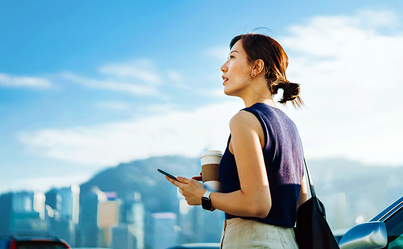 青空の下、スマートフォンとコーヒーカップを持った女性が遠くを見つめている。