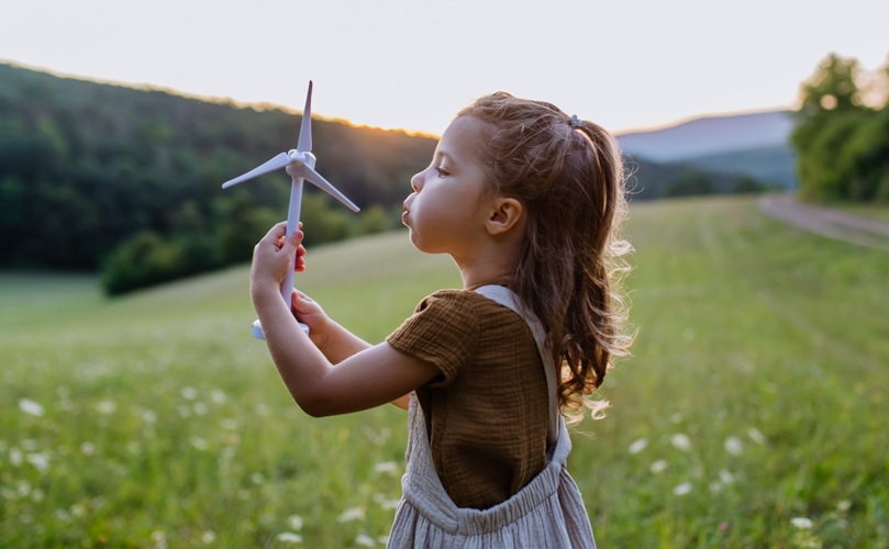 A little girl joyfully holds a colorful windmill in a sunny field, surrounded by tall grass and wildflowers.