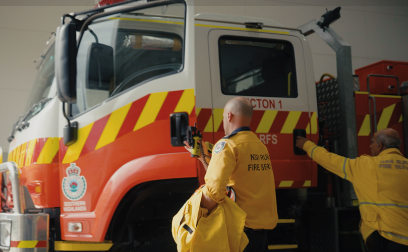Two men in yellow jackets stand beside a fire truck, ready for action in a community safety event.