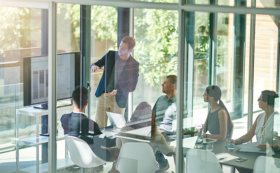 ガラス張りの会議室で、グラフを指しながらプレゼンテーションを行う男性