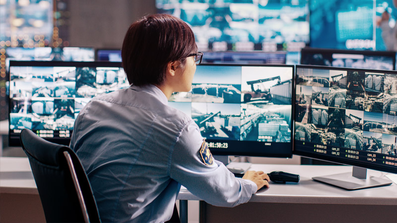 Univormuun pukeutunut henkilö istuu työpöydän ääressä ja katsoo kolmea näyttöä.