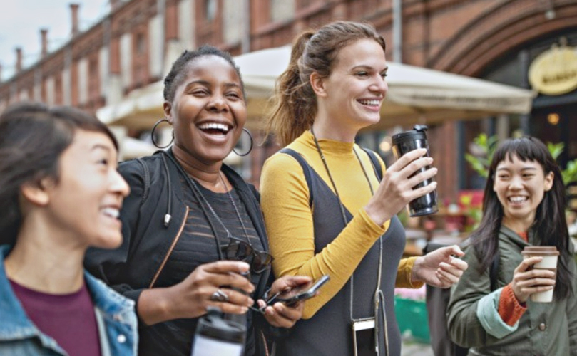 Một nhóm phụ nữ mỉm cười, đi xuống phố, trong khi đang cầm điện thoại và ly cà phê