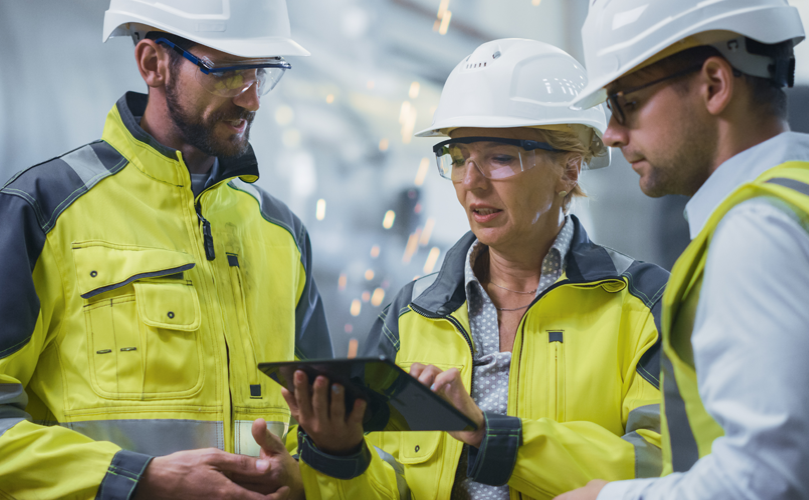 Three factory workers in hard hats and high-visibility safety jackets review information on an electronic handheld device while engaged in discussion