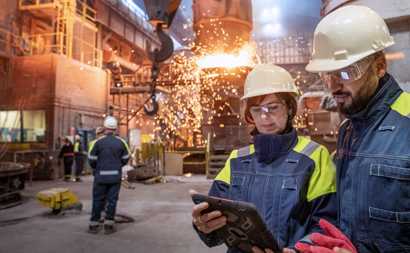 A man and woman in protective gear looking at a tablet in the middle of a factory