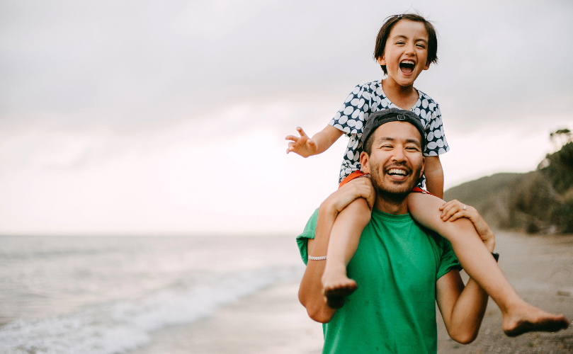 아이를 어깨에 메고 해변에 있는 남자, 둘 다 웃고 있습니다