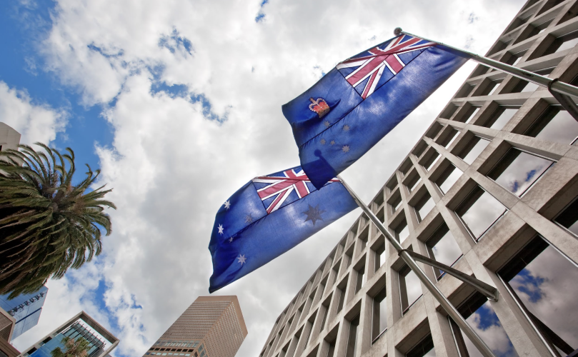 Shot taken from a low angle showcasing a building on the right, a palm tree on the left and two Australian flags in the middle