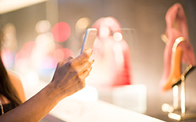 女性がスマートフォンのカメラで、ショーウィンドウ内の商品（ピンクのバッグや靴）を撮影している様子。