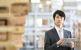 スーツを着たアジア系の男性が、倉庫でタブレットを持ち、遠くを見つめている様子。