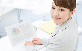 アジア系の若い女性が、白いオフィスでノートパソコンに向かい、笑顔で振り返っている様子。パソコンの画面にはグラフが表示されている。