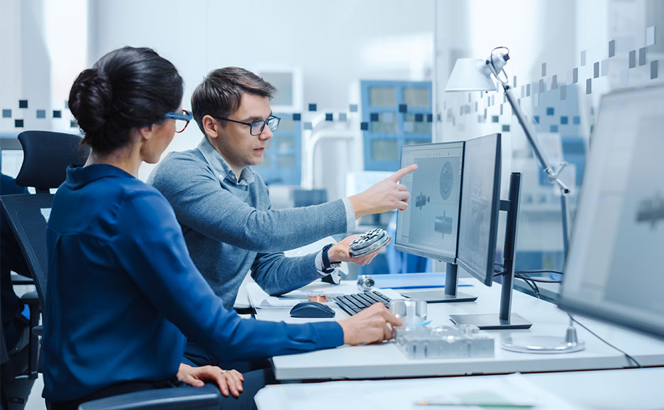 設計用デスクに並ぶ複数のモニター上で機械部品の3Dモデルやシミュレーション結果を表示し、操作デバイスを用いながら画面を指し示して設計内容を確認・議論する開発現場の様子