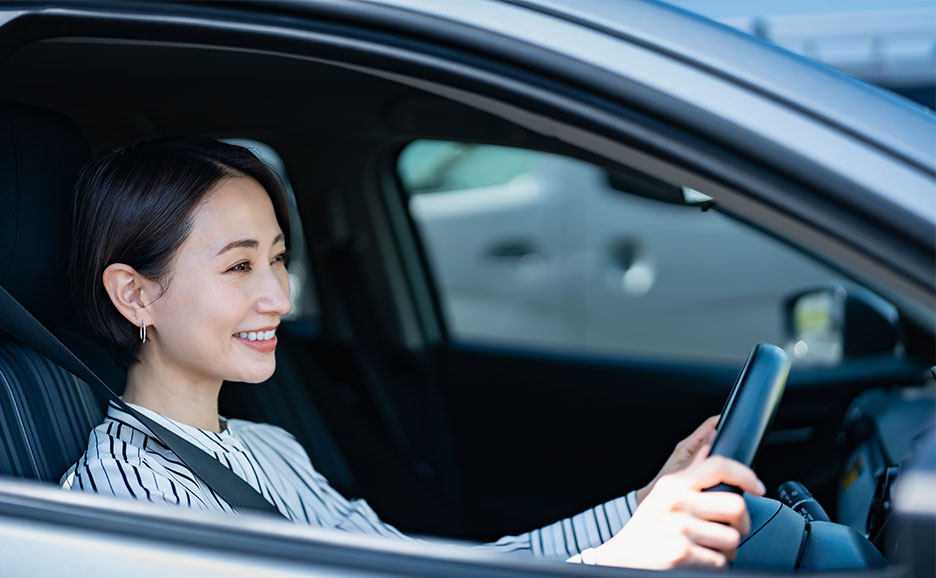 車の運転席でシートベルトを着用し、ハンドルを握って運転する人物を車外から横方向に撮影した写真。