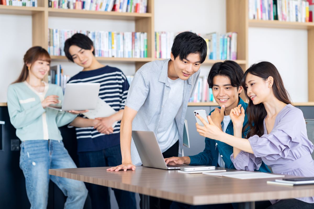 書籍が並んでいる部屋でのミーティング 手前3人はパソコンとスマートフォンを見ながら 奥の2人はノートパソコンを見ている