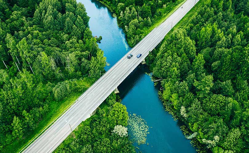 森林を流れる川に架かる橋を走る車の空撮風景。