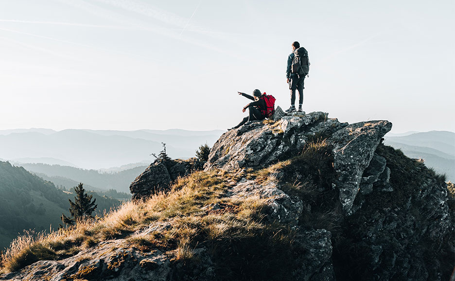 山頂で景色を眺める2人のハイカー。雄大な山々が広がる。