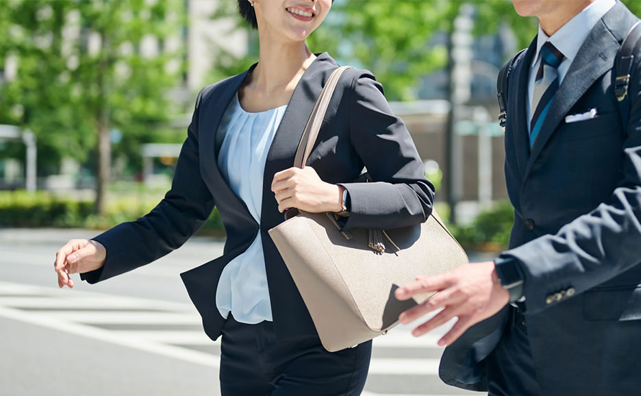 青いブラウスにスーツを着てベージュのバッグを持つ女性とスーツの男性が歩道で通り過ぎる瞬間。